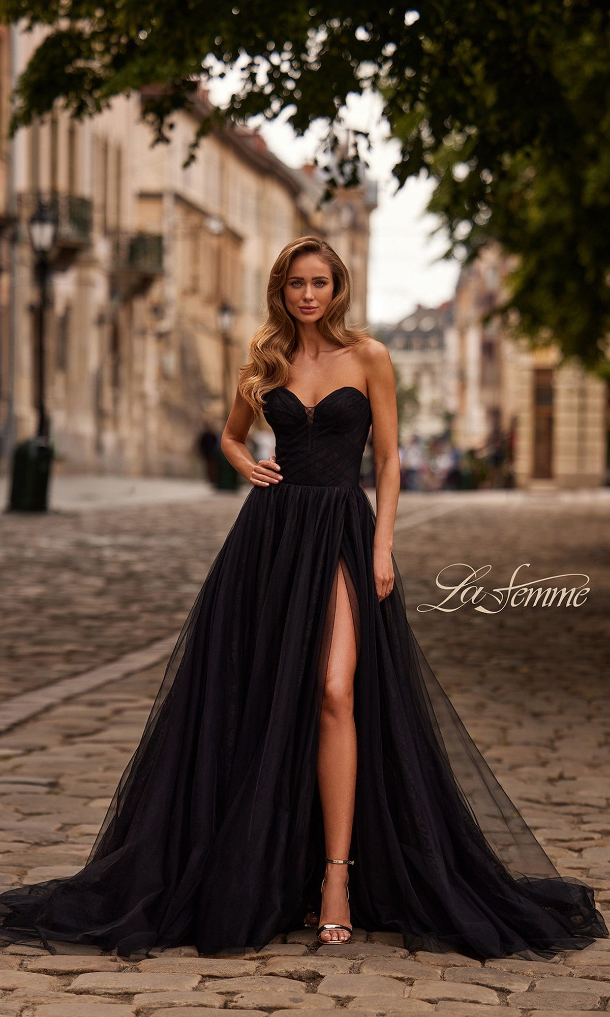 A woman with long wavy hair and heels stands on a cobblestone street in a black strapless high-slit gown, the La Femme 33307 Long Prom Dress. The
